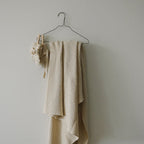 A knit heirloom blanket hanging on a white wall hook, with delicate lace trim and a woven pattern, primarily in a beige or light color.